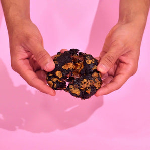 Hands holding a Crumbs & Doilies Caramel Cornflake New York Cookie with visible caramel and cornflake topping.